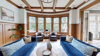 This distinguished two-story home blends classic architectural character with thoughtfully updated interiors. The grand entrance immediately impresses with gleaming hardwood floors, rich wood wainscoting, decorative columns, and a stately staircase with a patterned runner leading to the upper level. The main floor offers multiple living spaces, including a stunning rounded sitting room featuring leaded glass windows, coffered ceilings with warm wood detailing, and abundant natural light with lush outdoor views. A formal living room continues the elegant aesthetic with a marble fireplace, wood-beamed coffered ceilings, and wainscoting throughout. The combined dining room and kitchen area incorporates a striking natural stone accent wall, quartz countertops, and French doors that open to the outdoors. An additional living space with floor-to-ceiling French doors offers seamless garden views. Upstairs, the upper hallway is appointed with hardwood floors, a chandelier, and thoughtful built-in storage. The dedicated office features a fireplace and ample workspace. Two bathrooms serve the home, one of which includes a unique tanning bed and tile flooring, while the second features a classic multi-toned slate tile floor and vaulted ceiling detail. The home's generous scale across both floors provides abundant room for comfortable living.