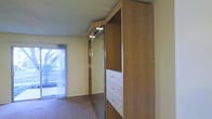 This single-floor space is highlighted by a prominent built-in wardrobe unit that extends nearly to the ceiling, combining practicality with thoughtful design. The unit features mirrored sliding doors that visually expand the room while offering convenient reflective functionality. Integrated white drawers provide organized storage, and open shelving on the adjacent panel adds versatile display or storage options. Recessed spotlights are mounted directly on the wardrobe unit, providing focused task lighting. The warm wood-toned cabinetry contrasts with the neutral cream-colored walls and textured popcorn ceiling. Soft, neutral carpeting in a muted brown tone covers the floor, contributing to a comfortable and understated ambiance. A sliding glass door fitted with sheer white curtains allows natural light to filter into the room, brightening the space and offering a view of outdoor greenery. The combination of mirrored surfaces, warm wood tones, and natural light creates a functional yet inviting atmosphere throughout this well-appointed space.