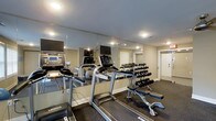 The fitness center at Fairway 28 is a dedicated, single-floor workout facility designed to support a well-rounded exercise routine. The space is outfitted with a selection of cardio equipment, including Landice treadmills and additional machines such as an elliptical and rowing equipment, each equipped with individual monitors for entertainment or workout tracking. A full dumbbell rack lines one wall, offering a wide range of free weights alongside a dedicated weight bench, making the space suitable for both strength and cardio training. Floor-to-ceiling mirrors span the length of one wall, visually expanding the room and allowing users to monitor their form during workouts. Recessed lighting illuminates the space evenly from above, creating a bright and functional atmosphere. The flooring is finished with durable rubber gym matting, providing both traction and cushioning underfoot. Neutral-toned walls and a clean, organized layout contribute to a focused and comfortable environment. A private door at the rear of the room suggests additional access or utility space. Overall, the fitness center offers a practical and well-maintained setting for residents seeking convenient on-site exercise amenities.