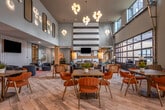 Located at 1399 Delashmut Ave in Columbus, Ohio, this generously sized single-floor community amenity space blends contemporary design with warm, eclectic details throughout. The entry foyer sets an immediate tone with its rich exposed brick wall, bold teal diagonal-paneled accent column, and light wood-look tile flooring that carries seamlessly through the entire space.

The main lounge area commands attention with soaring double-height ceilings and a dramatic floor-to-ceiling industrial-style glass garage door that floods the interior with natural light and provides open views to the exterior. Spherical globe pendant chandeliers hang overhead, complementing the airy, light-teal painted walls. Comfortable seating arrangements—including tufted leather sofas, curved lounge chairs, and eclectic area rugs—create distinct yet connected social zones.

A cozy sitting area features a linear fireplace framed by a bold black-and-white striped accent wall and a rustic brick feature wall with decorative open shelving. Adjacent is a billiards lounge anchored by a full-size pool table beneath an elegant multi-globe chandelier, with a bar-height counter along the wall for casual seating.

The kitchen area offers a fully equipped setup with a fluted wood island, quartz countertops, bar seating, stainless steel appliances, dark wood cabinetry, and vibrant pop-art-inspired wall murals that add an energetic, creative flair. Throughout, the space balances modern sophistication with inviting warmth.