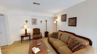 This single-floor residence offers a thoughtfully arranged layout with multiple living spaces designed for comfort and functionality. The living room serves as the heart of the home, featuring warm carpeting, a wood-framed sofa and matching armchair with patterned upholstery, a wooden coffee table, and a collection of framed artwork and decorative wall sconces that lend a classic, cozy character to the space. Floor lamps provide soft ambient lighting throughout.

The kitchen is equipped with warm oak cabinetry, a white electric range, ample counter space with light-toned countertops, a stainless steel sink, and tile flooring — offering a practical and efficient workspace. Adjacent to the kitchen, the dining area features a wooden dining table with white chairs beneath a multi-bulb chandelier, with drape-framed access to natural light.

The primary bedroom is generously sized with plush carpeting, a wooden bed frame with decorative ironwork, a matching dresser with mirror, a bedside table, and a walk-in closet. A second bedroom provides comfortable accommodations with its own closet, wooden furnishings, and soft carpet flooring. The full bathroom features a clean, white-tiled shower and tub combination with chrome fixtures, maintaining a simple and functional design throughout.