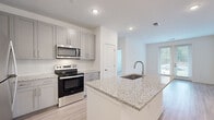 Located at 550 Webb Gin House Rd in Lawrenceville, GA, this single-floor residence offers a thoughtfully designed layout with contemporary finishes throughout. The open-concept kitchen serves as a central highlight, showcasing a large granite-topped island with an integrated sink, grey shaker-style cabinetry, a full suite of stainless steel appliances including a refrigerator, electric range, and over-the-range microwave, and a white subway tile backsplash. Light hardwood-style flooring flows through the main living areas, creating a bright and cohesive aesthetic. The living area is bathed in natural light through a set of French doors that open to outdoor views, adding both functionality and openness to the space. Bedrooms feature plush carpeting and large windows that invite additional natural light, with ceiling fans providing year-round comfort. The bathroom is finished with clean white tile surrounding a full soaking tub and shower combination, complemented by a grey vanity with stone countertops. Throughout the unit, crisp white walls, recessed lighting, and modern hardware reinforce a clean, move-in-ready atmosphere that is both practical and stylish.
