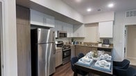 This single-floor residence showcases a thoughtfully designed interior with a clean, modern aesthetic throughout. The kitchen serves as a central highlight, featuring stainless steel appliances including a full-size refrigerator, gas range, and built-in microwave. Two-toned cabinetry — with upper white cabinets and lower warm wood-toned units — provides ample storage while contributing to a contemporary, layered look. Granite-style countertops tie the design together, complemented by a functional sink and additional prep space along the back wall. Recessed lighting illuminates the kitchen from above, enhancing the bright and open atmosphere created by the elevated ceiling height. Rich, dark hardwood-style flooring runs throughout, adding warmth and visual continuity. Adjacent to the kitchen, a dining area comfortably accommodates a full table and seating, making the space well-suited for both everyday meals and entertaining. The overall ambiance is polished and inviting, with a neutral color palette and modern finishes that create a cohesive, move-in-ready feel across the entire single-level layout.