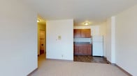 This single-floor space features an open layout that integrates the living area and kitchen into one cohesive zone. The living area is finished with neutral wall-to-wall carpeting, contributing to a warm and understated atmosphere. A partial dividing wall separates the living space from the kitchenette, which transitions to dark tile flooring. The kitchenette is equipped with warm-toned medium wood cabinetry offering ample upper and lower storage, dark laminate countertops, and a full-sized white refrigerator. Flush-mount ceiling light fixtures provide consistent overhead lighting throughout the space. The white walls keep the interior feeling bright and clean. A hallway visible from the main living area leads to additional spaces, including what appears to be a closet with a hanging rod. The layout is straightforward and functional, making efficient use of the available footprint. The transition between the carpeted living zone and the tiled kitchen area provides a clear visual distinction between the two functional spaces, contributing to an organized and practical overall design.