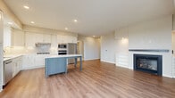 This well-appointed single-floor residence showcases a thoughtfully designed open-concept layout with consistent light wood flooring connecting the main living areas. The kitchen is a standout feature, offering white shaker cabinetry, quartz countertops, a subway tile backsplash, stainless steel appliances including a gas cooktop, wall oven, and refrigerator, along with a substantial dark-toned island providing generous prep and storage space. Large windows frame expansive outdoor views, filling the kitchen with natural light.

The living room is anchored by a fireplace flanked by built-in shelving on both sides, creating a refined focal point. Recessed lighting and neutral warm-toned walls contribute to a cohesive, inviting ambiance throughout.

The bedrooms feature plush carpeting and well-sized windows with elevated outdoor views. One bedroom includes access to a private balcony through sliding glass doors. A walk-in closet with wire shelving systems provides practical storage. The bathroom includes a glass-enclosed shower, tall built-in cabinetry, and light wood flooring. A dedicated laundry room houses a full-sized washer and dryer with overhead shelving for added convenience.