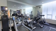 This fitness center is thoughtfully designed to provide a comprehensive workout environment within a compact, efficient layout. The space is outfitted with a selection of modern cardio equipment, including treadmills, elliptical trainers, and a recumbent bike, catering to a variety of fitness routines and training preferences. Full-length mirrored walls line one side of the room, visually expanding the space and allowing users to monitor their form during exercise. Recessed ceiling lights provide bright, even illumination throughout the facility, ensuring a well-lit environment at any time of day. Windows along the far wall allow natural light to filter into the space, contributing to an open and energizing atmosphere. The flooring features a dark, durable surface suited for fitness use, and the neutral-toned walls lend the room a clean, professional aesthetic. The equipment is arranged to allow comfortable movement between machines, making the space practical and easy to navigate. Overall, this fitness center offers a functional and inviting setting for cardiovascular training and general exercise.
