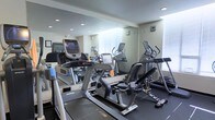 This fitness center is thoughtfully designed to provide a comprehensive workout environment within a compact, efficient layout. The space is outfitted with a selection of modern cardio equipment, including treadmills, elliptical trainers, and a recumbent bike, catering to a variety of fitness routines and training preferences. Full-length mirrored walls line one side of the room, visually expanding the space and allowing users to monitor their form during exercise. Recessed ceiling lights provide bright, even illumination throughout the facility, ensuring a well-lit environment at any time of day. Windows along the far wall allow natural light to filter into the space, contributing to an open and energizing atmosphere. The flooring features a dark, durable surface suited for fitness use, and the neutral-toned walls lend the room a clean, professional aesthetic. The equipment is arranged to allow comfortable movement between machines, making the space practical and easy to navigate. Overall, this fitness center offers a functional and inviting setting for cardiovascular training and general exercise.