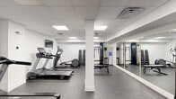 Located at 901 Leland St in Philadelphia's Francisville neighborhood, this fitness center offers a well-equipped workout facility within a compact, efficiently organized single-room layout. The space features a drop-panel ceiling with recessed flat lighting panels that provide bright, even illumination throughout, while crisp white walls and dark rubberized flooring create a clean, functional aesthetic.

The cardio zone is equipped with multiple Spirit treadmills and an elliptical trainer, each positioned near wall-mounted flat-screen TVs for entertainment during workouts. A dedicated spin bike station adds cycling options to the mix. For strength training, the room includes a multi-function cable machine with various attachment options, a dumbbell rack stocked with a full range of free weights, and a weight bench. A large wall-to-wall mirror with gold-trim framing spans one side of the room, visually expanding the space and allowing users to monitor their form.

Additional functional training accessories include a full rack of medicine balls and stability balls in various sizes, making the space suitable for dynamic and core-focused workouts. A water cooler is conveniently positioned for easy access. Structural columns punctuate the floor plan, and HVAC ceiling vents ensure proper climate control throughout. The overall design is straightforward and practical, prioritizing utility and ease of movement for a comprehensive fitness experience.
