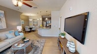 This single-floor residence offers a thoughtfully designed open-concept layout that seamlessly connects the living, dining, and kitchen areas. The living room is bright and welcoming, featuring light wood-look flooring, a ceiling fan, and sliding glass doors that lead to an outdoor patio, flooding the space with natural light. A dedicated media wall with a mounted flat-screen television anchors one side of the room, while a cozy seating arrangement completes the space.

The kitchen is a standout feature, boasting rich dark cabinetry, granite countertops, a curved breakfast bar peninsula, stainless steel appliances — including a refrigerator, microwave, and range — and pendant lighting overhead. It flows naturally into the dining area, which is highlighted by a round glass-top table, upholstered chairs, a globe pendant light, and a decorative gold-framed mirror.

The bedroom is a serene retreat, carpeted for comfort, with a large window that invites natural light and outdoor views. It offers ample space for a full furniture arrangement, complemented by a ceiling fan above.

The bathroom is clean and modern, featuring a walk-in glass-enclosed shower with built-in shelving and brushed nickel fixtures alongside open wall shelving for convenient storage. Throughout, the space maintains a cohesive, coastal-inspired aesthetic with neutral tones and tasteful décor accents.