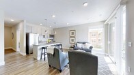 This two-floor space is designed with an open-concept layout that connects the kitchen, dining, and living areas into one cohesive, functional environment. The kitchen is outfitted with light-toned cabinetry, stainless steel appliances including a full-size refrigerator and microwave, and a sleek countertop island with bar seating — ideal for casual dining or meal preparation. Pendant lighting fixtures hang above the island, complementing the recessed lighting throughout the ceiling to provide layered illumination across the space.

The living area flows naturally from the kitchen, offering ample room for seating arrangements centered around a patterned area rug. A large window invites generous natural light into the space while providing views of the surrounding neighborhood. Neutral gray walls and white ceilings create a clean, contemporary backdrop that complements the warm-toned hardwood-style flooring running throughout the main level.

The overall aesthetic is modern and understated, combining practical design with a welcoming ambiance. The two-floor layout provides additional space beyond the main open-concept level, offering flexibility for varied living needs.