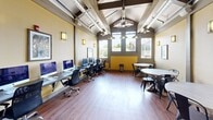 Located at 100 Hawk Ridge Dr in Iowa City, Iowa, this thoughtfully designed communal space combines industrial and contemporary elements to create a welcoming, multi-functional environment. The open floor plan is anchored by a striking vaulted ceiling with exposed wood beams and visible ductwork, lending an urban-loft aesthetic throughout. Warm hardwood-style flooring runs seamlessly across the entire space, complementing the golden-yellow accent walls and natural wood tones.

At the heart of the room, a floor-to-ceiling stone fireplace serves as a dramatic focal point, paired with a wall-mounted television and flanked by round dining tables with navy metal chairs — ideal for casual gatherings or collaborative meetings. Industrial-style pendant lighting hangs from the vaulted ceiling, casting a warm, inviting glow across the space.

A dedicated computer workstation area lines one wall, equipped with multiple desktop computers and ergonomic seating, making this space well-suited for productivity and remote work. Large windows along the perimeter invite abundant natural light and frame views of the surrounding landscape. An open balcony overlook with black metal railings extends the space outward, offering outdoor views and additional ventilation. Entry and exit are facilitated by a heavy-duty commercial door, reinforcing the space's shared-amenity character.