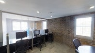 This single-floor workspace offers a thoughtfully designed environment that balances industrial aesthetics with practical office functionality. The most striking architectural feature is the exposed brick wall, which runs the length of the room and lends a warm, textured character to the space. The smooth white ceiling contrasts with the raw brickwork below, creating a visually dynamic interior.

The workspace is equipped with multiple workstations arranged along sleek black desks, each accompanied by ergonomic rolling chairs and desktop monitors, making it well-suited for collaborative or shared office use. Natural light filters in through windows fitted with horizontal blinds, keeping the space bright while allowing for light control.

A glass partition wall divides the area, adding a sense of openness and transparency while defining separate zones within the office. Recessed ceiling lights provide consistent overhead illumination throughout. The floor is finished with neutral stone-toned tiles that complement the overall palette of the room. Together, these elements create a professional yet character-rich workspace that balances modern office needs with an inviting, urban-industrial ambiance.