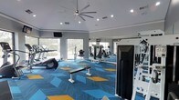 This single-story fitness center offers a thoughtfully designed workout environment that balances functionality with contemporary style. The space is anchored by vaulted ceilings that lend an open, airy feel, complemented by a large ceiling fan and recessed lighting that ensure comfortable conditions throughout. A wall of floor-to-ceiling windows along one side floods the interior with natural light while providing views of the surrounding landscape, creating an inviting atmosphere for exercise.

The flooring features a bold geometric carpet in shades of blue and gold, adding a dynamic visual energy to the space. Equipment is well-distributed, including treadmills and cardio machines positioned near the windows to take advantage of the natural light and views, alongside a selection of strength training machines and a flat bench for free weight exercises. Full-length mirrors along one wall visually expand the space while allowing users to monitor their form. Wall-mounted televisions provide entertainment options. The clean gray walls and modern finishes give the fitness center a polished, professional appearance suited to a variety of workout routines.