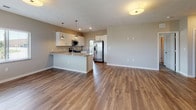 This single-floor residence presents a well-designed, open-concept layout with a cohesive neutral color palette of warm beige and gray tones throughout. The kitchen serves as a central highlight, featuring crisp white shaker-style cabinetry, speckled granite countertops, and a suite of stainless steel appliances including a side-by-side refrigerator with ice maker, electric range, and built-in microwave. A peninsula with an undermount sink and pendant lighting above provides additional prep space and separates the kitchen from the adjacent living area. Wood-look plank flooring extends throughout the main living and kitchen areas, while plush carpet covers the bedrooms. The open living space is bright and functional, with a flush-mount ceiling fixture and a window allowing natural light in. Bedrooms are generously proportioned, each featuring carpeted floors, windows with exterior views, and ceiling fans or overhead lighting. The primary bathroom includes a granite-topped vanity with white cabinetry, a large mirror, tile flooring, and a walk-in shower, with an adjacent walk-in closet providing ample storage. Textured ceilings and clean white trim are consistent throughout, contributing to a polished, move-in-ready aesthetic.