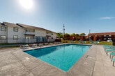 Located at 700 S Clinton St in Athens, Alabama, this outdoor pool area serves as a central amenity for the surrounding residential community. The centerpiece is a generously sized rectangular swimming pool with clear, blue water, designed to accommodate a large number of swimmers. Surrounding the pool is a wide concrete sundeck lined with numerous lounge chairs and seating arrangements, providing ample space for relaxation and sunbathing under the open Alabama sky. Colorful patio umbrellas are stationed throughout the deck, offering shaded seating areas with chairs and tables for casual outdoor gatherings. The pool area is fully enclosed by a black iron perimeter fence, ensuring security and controlled access. A dedicated pool house building is situated at one end of the space, constructed with vinyl siding and featuring teal-painted entry doors. Posted pool rules and safety equipment, including a life ring, are visibly displayed on the exterior of the pool house. The surrounding landscape includes mature trees that provide a natural backdrop, complementing the open, sun-filled atmosphere. The multi-story residential buildings of the community are visible beyond the fencing, reinforcing the pool area's role as a shared outdoor hub for residents.