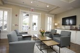 This single-floor communal lounge is thoughtfully designed to balance comfort and functionality within a refined, traditional aesthetic. The space features warm hardwood flooring throughout, complemented by neutral warm-toned walls, crown molding, and tray ceiling details that lend an air of architectural polish.

A central seating area anchors the room, furnished with upholstered blue-gray armchairs arranged around a coffee table, creating an inviting gathering space. A dedicated refreshment station lines one wall, featuring white cabinetry with granite countertops, a coffee maker, and a wall-mounted flat-screen television above—ideal for casual interaction and relaxation.

Along an adjacent wall, a built-in workstation accommodates multiple computer terminals and a printer, providing a functional business center area. A large framed cityscape photograph adds a decorative focal point nearby.

Natural light fills the space through plantation-shuttered windows and a set of glass-paneled French doors, which open directly to an outdoor area, offering a seamless indoor-outdoor connection. A glimpse through interior glass sliding doors reveals a connected lobby or common area with columns and a staircase, underscoring the broader scope of the building. Recessed lighting and wall sconces complete the warm, welcoming ambiance throughout.