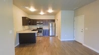 This single-floor space showcases an open-concept layout that seamlessly connects the kitchen and living areas, creating a functional and inviting environment. The kitchen is well-appointed with dark espresso-toned cabinetry that provides ample storage, complemented by light granite-style countertops that offer a pleasing contrast. Stainless steel appliances, including a refrigerator, electric range, microwave, and dishwasher, lend a modern, cohesive aesthetic to the cooking space. A kitchen island or peninsula extends the counter space, making it ideal for meal preparation or casual dining.

Warm hardwood-style flooring flows continuously throughout the open area, adding both visual warmth and durability underfoot. The neutral-toned walls and flush-mount ceiling lighting create a bright, clean atmosphere. The open layout between the kitchen and the adjoining living space allows for flexible furniture arrangements and a sense of generous, uninterrupted square footage. Additional doors visible from the main area suggest access to further rooms, such as bedrooms or a hallway, contributing to the overall functionality of this well-designed single-level space.