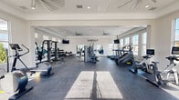 The Yardly fitness center is a generously sized, single-floor gym facility designed to support a comprehensive workout experience. The space features a clean, modern aesthetic with bright white walls, a tray ceiling with recessed lighting, and durable rubber-speckled flooring throughout that is both functional and resilient underfoot.

Natural light pours in abundantly through large windows positioned along multiple walls, including a row of clerestory windows near the ceiling that further enhance the open, airy atmosphere. Ceiling fans are installed throughout the space to promote air circulation, while wall-mounted flat-screen TVs provide entertainment during workouts.

The cardio zone is well-equipped with multiple treadmills, stair climbers, elliptical trainers, and rowing machines, all arranged to maximize space and usability. The strength training area features a multi-station cable machine, a power rack with an adjustable bench, and an extensive dumbbell rack covering a wide range of weights. Full-length mirrors line one wall of the strength area, allowing users to monitor their form effectively.

A glass-panel entry door offers views of the surrounding community and allows additional natural light to flow into the space. The layout is thoughtfully organized, ensuring ample room to move freely between equipment zones, making this fitness center a well-rounded amenity for residents.
