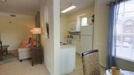 This single-floor space is characterized by a clean, functional layout with a warm, inviting ambiance. The kitchen is a standout feature, outfitted with white cabinetry, a full-size refrigerator, electric range, and built-in microwave. A mosaic tile backsplash adds a subtle decorative touch against the neutral cream-toned walls. The kitchen receives natural light through a window above the counter, keeping the space bright and airy. A built-in overhead light fixture provides additional illumination.

A half-wall partition separates the kitchen from the adjacent dining and living areas, creating a sense of openness while maintaining distinct zones within the layout. The transition between spaces is smooth, with light-toned flooring running consistently throughout. A woven dining chair is partially visible near the kitchen entrance, hinting at a cozy dining nook nearby.

The living area, glimpsed through the arched passageway, features warm lamplight, a wooden accent console, and tasteful wall art, contributing to the space's comfortable and well-appointed character. Crown molding details along the upper walls add a refined architectural touch throughout.