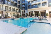 Located in Sandy Springs, Georgia, this stunning courtyard amenity space is designed to deliver a resort-like experience within a modern residential community. The architecture combines classic light-toned brick exteriors with contemporary dark metal balcony railings and expansive floor-to-ceiling glass panels, creating a refined aesthetic that frames the outdoor living area on all sides.

At the heart of the space is a generously sized swimming pool with crystal-clear blue water, complemented by an adjacent spa and a raised water feature with a stone surround that adds a sophisticated focal point. The spacious pool deck is finished in smooth concrete pavers, providing ample room for relaxation and social gatherings. Rows of sleek, modern chaise lounge chairs line the sun deck, offering residents comfortable spaces to unwind.

Lush landscaping, including mature magnolia trees, ornamental shrubs, and neatly mulched garden beds, softens the hardscape and adds natural beauty throughout the courtyard. A wooden pergola provides a shaded gathering spot near the water's edge, enhancing the outdoor living experience. Large glass-panel entry doors connect the courtyard to the building's interior, ensuring seamless indoor-outdoor flow. The entire space is enclosed by the multi-story residential building, creating an intimate and private atmosphere ideal for relaxation and community living.