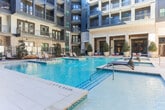 Located in Sandy Springs, Georgia, this stunning courtyard amenity space is designed to deliver a resort-like experience within a modern residential community. The architecture combines classic light-toned brick exteriors with contemporary dark metal balcony railings and expansive floor-to-ceiling glass panels, creating a refined aesthetic that frames the outdoor living area on all sides.

At the heart of the space is a generously sized swimming pool with crystal-clear blue water, complemented by an adjacent spa and a raised water feature with a stone surround that adds a sophisticated focal point. The spacious pool deck is finished in smooth concrete pavers, providing ample room for relaxation and social gatherings. Rows of sleek, modern chaise lounge chairs line the sun deck, offering residents comfortable spaces to unwind.

Lush landscaping, including mature magnolia trees, ornamental shrubs, and neatly mulched garden beds, softens the hardscape and adds natural beauty throughout the courtyard. A wooden pergola provides a shaded gathering spot near the water's edge, enhancing the outdoor living experience. Large glass-panel entry doors connect the courtyard to the building's interior, ensuring seamless indoor-outdoor flow. The entire space is enclosed by the multi-story residential building, creating an intimate and private atmosphere ideal for relaxation and community living.