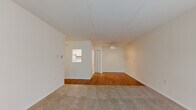 This single-floor residential unit offers a practical and functional layout with a clean, neutral aesthetic throughout. The main living area is generously proportioned, featuring a combination of warm hardwood-style flooring and plush carpet, creating distinct zones within the open space. Large vertical blinds frame a sliding glass door that opens to a balcony, allowing ample natural light to fill the room. A ceiling fan with built-in lighting provides both comfort and illumination.

The galley-style kitchen is efficiently designed with white cabinetry accented by warm wood trim, light countertops, hardwood-style flooring, and includes a range and dishwasher. A pass-through window connects the kitchen to the living area, enhancing the sense of openness.

The bedroom is a comfortable, carpeted room featuring a window with exterior greenery views, a closet with bifold doors, and a baseboard heating unit. The full bathroom is finished with white square tile surrounding the bathtub and shower combo, complemented by light tile flooring, maintaining a bright and clean appearance throughout. The overall ambiance of the unit is understated and move-in ready, with a neutral color palette that offers a versatile backdrop for any style of furnishing.