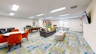 This generously sized game and recreation room offers a lively, multi-functional space designed for social engagement and leisure. The open floor plan is unified by a striking mosaic-patterned carpet in shades of teal, yellow-green, and gray, which anchors the room with vibrant energy and a modern aesthetic.

At the heart of the space, a full-size pool table takes center stage alongside a foosball table, providing classic recreational options. A ping-pong table occupies its own dedicated zone near a window that allows natural light to fill the room. A lounge area features a plush gray sofa, patterned ottoman stools, and a wall-mounted flat-screen TV set against a warm wood-paneled accent wall, creating a comfortable media viewing nook.

A separate seating vignette with a round white table and bold orange upholstered chairs offers an inviting spot for casual conversation or tabletop games, accented by a triptych of framed photographic artwork on the wall behind. Vending machines positioned near the entrance add convenience for snacks and beverages.

Recessed linear LED lighting fixtures provide bright, even illumination throughout, while windows along one wall bring in views of the surrounding outdoor greenery. The overall ambiance is energetic, colorful, and welcoming, making this a well-appointed communal destination for recreation and social interaction.