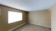 This 1-bedroom, 1-bathroom unit is laid out across a single floor, offering a straightforward and functional living arrangement. The bedroom pictured features wall-to-wall carpet that adds warmth underfoot, paired with neutral tan walls that provide a versatile backdrop for a variety of décor styles. A large window with horizontal blinds allows natural light to fill the room, contributing to a bright and open feel. A baseboard heating unit runs along the exterior wall, ensuring reliable warmth during cooler months. The textured popcorn ceiling adds a classic finish consistent throughout the unit. White trim and baseboards provide clean contrast against the neutral tones of the walls and flooring, lending a tidy, cohesive appearance. The room offers ample open space, making it adaptable for various furniture configurations. Overall, the unit presents a clean, no-frills living environment well-suited for individuals or couples seeking a comfortable and manageable space with essential features already in place.