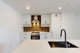 Located at 1330 S Fair St in Arlington, VA, this thoughtfully designed one-bedroom, one-bathroom residence offers a clean, contemporary aesthetic throughout. Light wood-look flooring flows seamlessly across the open-concept living area, connecting the living room, dining space, and kitchen into a cohesive and functional layout. The kitchen is a standout feature, showcasing flat-front white cabinetry with matte black hardware, a warm wood-toned backsplash, stainless steel range with a sleek hood vent, and a large center island with a white quartz countertop, undermount sink, and dark cabinetry base. Natural light pours in through floor-to-ceiling sliding glass doors that open to a private balcony, offering open views of the Arlington skyline. The bedroom is a bright, comfortable retreat with dual windows framing urban views and includes a built-in Murphy bed cabinet. Two closets provide generous storage, including a walk-in closet outfitted with adjustable shelving and hanging rods. The bathroom features tile flooring, a sleek dark-toned vanity with integrated sink, and direct access to the walk-in closet. In-unit laundry adds everyday convenience. Clean white walls and recessed lighting throughout contribute to a bright, modern ambiance.