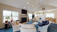 This inviting single-floor space exudes a relaxed coastal charm throughout its thoughtfully designed layout. The main living area is anchored by a striking stone fireplace with a mounted flat-screen television above, flanked by large windows that flood the room with natural light and offer views of the surrounding greenery. Vaulted ceilings with recessed lighting and a ceiling fan add an airy, open feel, while rich blue carpeting and a mix of neutral and navy furnishings reinforce the nautical theme. Decorative touches such as a ship's wheel and sailboat artwork enhance the seaside atmosphere throughout.

A secondary lounge area connects via elegant French doors, providing a versatile retreat with additional seating, vaulted ceilings, and direct access to the outdoors through a set of double French doors framed by ample windows. The dining area accommodates generous gatherings around a large wooden table with seating for eight, brightened by a wide window and complemented by sailing-themed wall art. The kitchen is clean and functional, featuring white cabinetry, full appliances including a range, microwave, and refrigerator, a double sink, and a window overlooking the outdoors — offering both practicality and a bright, welcoming workspace.