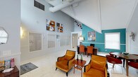This well-appointed single-floor space makes a striking first impression with its dramatic vaulted ceilings, exposed white beams, and visible ductwork that lend an industrial-chic character to the interior. The layout is open and inviting, seamlessly blending a comfortable lounge area with a functional workspace. The lounge features plush amber-toned armchairs arranged around a sleek wooden coffee table, creating an ideal setting for relaxed conversation or informal meetings. A bold teal accent wall anchors one side of the space, adding a vibrant pop of color that contrasts beautifully with the cool light-grey walls throughout. The workspace area is defined by a dark wood desk and warm orange chairs, complemented by a framed piece of wall art that adds visual interest. Contemporary abstract paintings adorn the walls, contributing to the space's modern, curated aesthetic. Light-toned tile flooring runs throughout, enhancing the sense of openness and reflecting the abundant natural light that filters in through multiple windows dressed with white horizontal blinds. A ceiling fan with a pendant light fixture adds both function and style overhead. The overall ambiance is energetic yet comfortable, blending bold color choices with clean architectural lines.