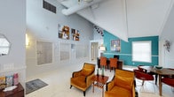 This well-appointed single-floor space makes a striking first impression with its dramatic vaulted ceilings, exposed white beams, and visible ductwork that lend an industrial-chic character to the interior. The layout is open and inviting, seamlessly blending a comfortable lounge area with a functional workspace. The lounge features plush amber-toned armchairs arranged around a sleek wooden coffee table, creating an ideal setting for relaxed conversation or informal meetings. A bold teal accent wall anchors one side of the space, adding a vibrant pop of color that contrasts beautifully with the cool light-grey walls throughout. The workspace area is defined by a dark wood desk and warm orange chairs, complemented by a framed piece of wall art that adds visual interest. Contemporary abstract paintings adorn the walls, contributing to the space's modern, curated aesthetic. Light-toned tile flooring runs throughout, enhancing the sense of openness and reflecting the abundant natural light that filters in through multiple windows dressed with white horizontal blinds. A ceiling fan with a pendant light fixture adds both function and style overhead. The overall ambiance is energetic yet comfortable, blending bold color choices with clean architectural lines.