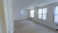 This single-floor space offers a clean and functional layout with a neutral, move-in-ready aesthetic. The main living area is generously proportioned, featuring wall-to-wall carpeting in a light beige tone that adds warmth and comfort underfoot. Three large windows dressed with vertical blinds line the walls, allowing abundant natural light to fill the room and creating a bright, airy atmosphere throughout the day. A radiator beneath one of the windows provides an efficient heating source, characteristic of classic residential construction. The smooth white walls and textured ceiling contribute to a simple, understated interior that offers a versatile canvas for personalization. The open layout of the living area promotes a spacious feel, with ample room for a variety of furniture arrangements. The overall ambiance is calm and welcoming, with the natural light and neutral palette working together to create an inviting environment. This space is well-suited for those seeking a straightforward, comfortable single-floor living arrangement with practical features and a timeless interior design.