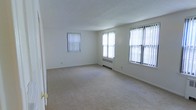 This single-floor space offers a clean and functional layout with a neutral, move-in-ready aesthetic. The main living area is generously proportioned, featuring wall-to-wall carpeting in a light beige tone that adds warmth and comfort underfoot. Three large windows dressed with vertical blinds line the walls, allowing abundant natural light to fill the room and creating a bright, airy atmosphere throughout the day. A radiator beneath one of the windows provides an efficient heating source, characteristic of classic residential construction. The smooth white walls and textured ceiling contribute to a simple, understated interior that offers a versatile canvas for personalization. The open layout of the living area promotes a spacious feel, with ample room for a variety of furniture arrangements. The overall ambiance is calm and welcoming, with the natural light and neutral palette working together to create an inviting environment. This space is well-suited for those seeking a straightforward, comfortable single-floor living arrangement with practical features and a timeless interior design.