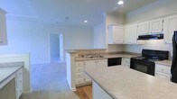 This single-story space showcases an open-concept layout that connects the kitchen to the surrounding living areas, creating a cohesive and functional flow throughout. The kitchen is outfitted with crisp white cabinetry offering generous storage through both upper and lower cabinets, complemented by neutral-toned laminate countertops that extend across a peninsula with an integrated double sink. The peninsula serves as a natural divider between the kitchen and the adjoining carpeted living space, making it ideal for both meal preparation and casual interaction. Black appliances, including a range and dishwasher, provide a subtle contrast against the white cabinetry. Recessed lighting in the kitchen ceiling ensures even, practical illumination throughout the space. The transition from tile or hardwood-style flooring in the kitchen to plush carpeting in the living area adds textural variety underfoot. Neutral wall tones throughout lend the space a clean, versatile aesthetic. A hallway visible in the background suggests additional rooms beyond, indicating a well-organized floor plan designed for comfortable everyday living.