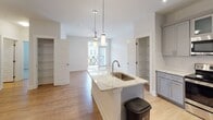 Located at 1000 Fazio Dr in Pinehurst, North Carolina, this single-floor residence offers a refined blend of modern design and functional living. The open-concept layout is anchored by a sleek kitchen featuring gray shaker-style cabinetry, granite countertops with subtle veining, a white subway tile backsplash, and stainless steel appliances including a refrigerator, range, and microwave. A large central island with an undermount sink and pendant lighting serves as both a prep space and a natural gathering point, flowing seamlessly into the living area, which is bathed in natural light from oversized windows with a balcony view beyond. Warm hardwood-style flooring runs throughout the main living and kitchen areas, lending continuity and warmth to the space. The bedroom is generously proportioned, finished with plush carpet, dual windows with blinds, and a ceiling fan for year-round comfort. The bathroom is well-equipped with gray cabinetry, marble-style countertops, a large framed mirror, and a glass-enclosed walk-in shower. A dedicated laundry area with a full-size washer and dryer adds everyday convenience. Multiple closets and built-in storage throughout the residence enhance its practical appeal, making this Pinehurst home as functional as it is stylish.