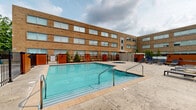 Located on Brookpark Road in Fairview Park, Ohio, this outdoor recreational area offers a well-appointed pool deck designed for relaxation and leisure. The centerpiece is a generously sized rectangular swimming pool with clear, inviting turquoise water, complemented by a circular whirlpool spa positioned nearby on the stamped concrete surround. The pool deck is enclosed by a sturdy black iron fence with warm-toned wood-accented gate panels, providing both security and aesthetic appeal. A collection of cushioned lounge chairs lines the perimeter of the deck, offering ample space for sunbathing and outdoor relaxation. The surrounding multi-story building features a classic tan brick facade with large, evenly spaced windows that overlook the pool area, creating a cohesive and well-maintained atmosphere. Mature trees visible beyond the enclosure add a touch of natural greenery to the setting. The overall layout is thoughtfully arranged, with easy access points and seating areas that make the space functional for both individual guests and groups. The open-sky setting allows for abundant natural light throughout the day, enhancing the overall appeal of this outdoor amenity.