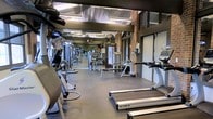 This fitness center offers a comprehensive workout environment designed to accommodate a variety of exercise needs. The space is defined by its industrial aesthetic, featuring exposed brick walls, structural columns, and a high ceiling with corrugated metal panels and visible ductwork that give the room an open, airy feel. Overhead fluorescent lighting ensures the entire floor is well-lit throughout.

The gym floor is finished with durable rubber flooring, providing both safety and comfort for users. A generous selection of cardio equipment is prominently featured, including TRUE-brand treadmills and stationary bikes, positioned for easy access. The strength training area is well-stocked with a variety of cable machines, weight benches, and multi-use resistance equipment arranged to allow ample space between stations.

Large windows and glass-paneled doors along one wall allow natural light to filter into the space, contributing to an inviting atmosphere. Full-length mirrors are strategically placed to help users monitor their form. The layout is thoughtfully organized, creating a smooth flow between cardio and strength zones. Overall, this fitness center combines functional design with a robust equipment offering to support a wide range of fitness goals.