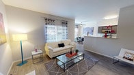This single-story space showcases an open-concept design that integrates the living, dining, and kitchen areas into one cohesive and functional layout. The living area serves as the central hub, featuring wood-look flooring underfoot and a neutral color palette on the walls that creates a calm, welcoming atmosphere. A large window dressed with patterned curtains allows natural light to fill the space, enhancing its airy feel. A glass-top coffee table and area rug anchor the seating area, adding contemporary style.

The space flows naturally into a dedicated dining area, where a ceiling fan overhead provides both practical ventilation and a decorative element. Beyond the dining area, a raised kitchen bar counter provides a visual boundary while maintaining the open connection between spaces. The kitchen features dark cabinetry and lighter countertops, offering a contrast that adds depth to the overall aesthetic.

Throughout the space, consistent wood-look flooring ties each zone together, while the neutral tones on the walls allow the furnishings and décor accents to stand out. The overall ambiance is modern, bright, and well-organized, making it a comfortable and versatile living environment.
