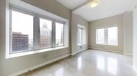 This single-floor space offers a clean, open layout with a polished terrazzo floor that reflects light throughout the interior, creating a bright and airy atmosphere. The neutral, warm-toned walls provide a versatile backdrop suited to a variety of uses. Large, deep-set windows dominate the main area, flooding the space with abundant natural light and framing impressive views of the surrounding urban skyline, including prominent multi-story buildings that speak to the elevated position of this space within the city. Additional windows along the adjoining wall further enhance the sense of openness and ensure consistent daylight from multiple angles. The elevated ceilings contribute to the generous sense of volume throughout, making the space feel expansive and unencumbered. Architectural details such as the deep window sills and clean wall transitions add a touch of structural character without detracting from the minimalist aesthetic. The overall ambiance is bright, modern, and functional, well-suited for use as a professional office, creative studio, or flexible commercial space. The combination of polished floors, sweeping city views, and abundant natural light makes this a distinctive and visually compelling environment.