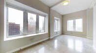 This single-floor space offers a clean, open layout with a polished terrazzo floor that reflects light throughout the interior, creating a bright and airy atmosphere. The neutral, warm-toned walls provide a versatile backdrop suited to a variety of uses. Large, deep-set windows dominate the main area, flooding the space with abundant natural light and framing impressive views of the surrounding urban skyline, including prominent multi-story buildings that speak to the elevated position of this space within the city. Additional windows along the adjoining wall further enhance the sense of openness and ensure consistent daylight from multiple angles. The elevated ceilings contribute to the generous sense of volume throughout, making the space feel expansive and unencumbered. Architectural details such as the deep window sills and clean wall transitions add a touch of structural character without detracting from the minimalist aesthetic. The overall ambiance is bright, modern, and functional, well-suited for use as a professional office, creative studio, or flexible commercial space. The combination of polished floors, sweeping city views, and abundant natural light makes this a distinctive and visually compelling environment.