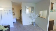 This single-floor residence presents a clean, understated aesthetic defined by neutral wall tones and wall-to-wall carpeting throughout. The interior layout features an open transitional area that connects the main living space to the bedroom, creating a sense of continuity and ease of movement. A partial dividing wall serves as a design focal point, adorned with a silver-framed mirror that reflects light and adds visual depth to the space. A decorative white branch arrangement in a modern vase adds an organic touch to the otherwise minimal décor. A second mirror is mounted on an adjacent wall above a small wooden ledge, providing a functional yet stylish accent. The bedroom, visible through the open passageway, is carpeted and bright, with a framed wall print adding a pop of color. The hallway beyond leads to additional areas of the unit. The overall ambiance is calm and livable, with soft lighting and muted tones contributing to a relaxed atmosphere. The layout makes efficient use of the available space while maintaining an open, airy feel throughout.