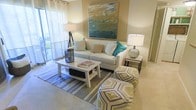This single-floor space centers around a well-appointed living area designed with a clean, contemporary aesthetic. The living room is finished in warm neutral tones, with carpeted flooring that adds a comfortable, inviting feel underfoot. A large, light-colored sofa serves as the focal point of the seating arrangement, dressed with teal and natural-toned accent pillows that complement the room's color palette. A bold abstract canvas in shades of teal, coral, and gold anchors the wall above the sofa, adding an artistic element to the space.

The room is furnished with a white wooden coffee table, offering both surface space and lower shelf storage, set atop a blue and white striped area rug. Geometric-patterned ottomans in gold and grey tones provide flexible additional seating. Flanking the sofa, coordinating table lamps in tripod and ceramic styles contribute warm ambient lighting.

Natural light filters into the space through sheer curtains along a sliding glass door, which opens to a private outdoor balcony or patio area, extending the living space beyond its interior. Overall, the space presents a cohesive, relaxed yet polished atmosphere.