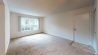 Located at 5820 Murray Dr, Unit B19, in Hanahan, South Carolina, this single-floor residence offers a straightforward and functional layout with well-defined living spaces throughout. The living area is generously sized, featuring neutral-toned carpet, a ceiling fan with light fixture, and a large window dressed with vertical blinds that allows abundant natural light to fill the room. A closet and entry door are conveniently positioned near the front of the living space. Adjacent to the living area, the kitchen is equipped with warm wood cabinetry, a refrigerator, and a louvered pantry or utility door, offering practical storage and workspace. The bedroom is a comfortable, well-proportioned room with plush carpet, a window with blinds providing views of the surrounding greenery, a textured ceiling, and a sliding closet for ample storage. The full bathroom is cleanly appointed with tile flooring, a white tiled tub and shower surround, a toilet, and a vanity featuring a warm wood cabinet base, white countertop, and a mirror above. Throughout the unit, the neutral color palette and consistent finishes create a cohesive and move-in-ready environment suited for everyday living.