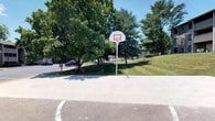 This outdoor recreational space features a well-maintained concrete basketball court surrounded by a lush, green landscape. The court is equipped with a standard regulation-height basketball hoop mounted on a sturdy post, complete with a white backboard and orange rim. Court boundary lines are clearly marked on the smooth concrete surface, providing a defined playing area for players of all skill levels.

The space is framed by a generous canopy of mature deciduous and evergreen trees, offering natural shade along the perimeter and contributing to a pleasant, park-like atmosphere. A gently sloping grassy hill borders one side of the court, adding to the open, airy feel of the environment. The surrounding grounds are neatly manicured, with well-kept lawns enhancing the overall appeal.

Under bright, sunny skies, the space is bathed in abundant natural light, making it inviting for daytime recreational activities. A parking area is visible nearby, providing convenient access for visitors. The combination of open space, mature tree coverage, and quality court facilities makes this an excellent venue for casual pickup games, organized play, or simply enjoying the outdoors.