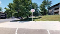 This outdoor recreational space features a well-maintained concrete basketball court surrounded by a lush, green landscape. The court is equipped with a standard regulation-height basketball hoop mounted on a sturdy post, complete with a white backboard and orange rim. Court boundary lines are clearly marked on the smooth concrete surface, providing a defined playing area for players of all skill levels.

The space is framed by a generous canopy of mature deciduous and evergreen trees, offering natural shade along the perimeter and contributing to a pleasant, park-like atmosphere. A gently sloping grassy hill borders one side of the court, adding to the open, airy feel of the environment. The surrounding grounds are neatly manicured, with well-kept lawns enhancing the overall appeal.

Under bright, sunny skies, the space is bathed in abundant natural light, making it inviting for daytime recreational activities. A parking area is visible nearby, providing convenient access for visitors. The combination of open space, mature tree coverage, and quality court facilities makes this an excellent venue for casual pickup games, organized play, or simply enjoying the outdoors.