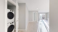 This thoughtfully designed 3-bedroom, 2-bathroom single-floor unit offers a clean, contemporary aesthetic throughout. Light-toned hardwood flooring and soft grey walls create a bright, cohesive atmosphere across all rooms. The modern kitchen is outfitted with high-gloss white cabinetry extending to the ceiling, stone countertops, stainless steel appliances, and recessed lighting, providing a polished and functional cooking space. The open living area is bathed in natural light from large sliding glass doors that lead to a private balcony overlooking a landscaped outdoor setting. A ductless mini-split system in the living area ensures year-round climate comfort. The bedrooms are well-proportioned, each featuring light hardwood floors, neutral grey walls, and windows that invite natural light; one bedroom includes a closet and a baseboard heater. The primary bedroom offers direct outdoor access. Two bathrooms serve the unit — one featuring a full soaker tub with a shower surround and a modern vanity, and the other offering a sleek walk-in glass shower with a white vanity. A dedicated in-unit laundry closet with a stacked washer and dryer adds everyday convenience. The overall design is modern, minimal, and move-in ready.