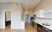 This single-floor unit showcases a contemporary design with light hardwood flooring, neutral gray walls, and recessed lighting throughout. The kitchen is well-appointed with a two-toned cabinet scheme pairing dark lower cabinets against white upper cabinets, complemented by light countertops, a subway tile backsplash, and stainless steel appliances including a built-in microwave and dishwasher. A built-in countertop ledge along one wall provides additional workspace or dining space. The open layout allows the kitchen to flow seamlessly into the living area, which features a built-in Murphy bed system with integrated shelving and cabinetry, offering flexible use of the space. Large floor-to-ceiling sliding glass doors flood the living area with natural light and open onto a private balcony with views of the surrounding neighborhood. The bathroom is clean and functional, featuring a dark-cabinet vanity with a white countertop, a full soaking tub with shower, and a stacked washer and dryer unit conveniently tucked within the same space. The entryway provides a smooth transition into the unit with tall ceilings enhancing the overall sense of openness.
