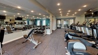 This fitness center is thoughtfully designed to accommodate a wide range of workout routines within a single, open-plan space. The interior is defined by bold teal accent walls contrasted against light gray surfaces, creating an energetic yet clean atmosphere. Recessed ceiling lighting illuminates the entire floor evenly, while multiple windows along the teal-painted wall invite natural light and views of surrounding greenery, enhancing the workout environment.

The space is organized into distinct functional zones. A dedicated cardio area houses multiple treadmills, elliptical trainers, and a stationary bike, all positioned in front of a large wall-mounted mirror and a flat-screen television. An adjoining free weights section features a full rack of dumbbells alongside an adjustable bench, supported by floor-to-ceiling mirrors that amplify the sense of space. The functional training zone includes a cable machine, a wall-mounted mat for stretching or bodyweight exercises, a kettlebell rack, and additional accessories for varied training.

Throughout the space, large-format rubber tile flooring provides durability and impact absorption. Glass doors connect to an additional cardio area, ensuring the layout remains open and well-ventilated. The overall design balances practicality with a modern, motivating aesthetic.
