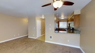 This two-floor residence offers a comfortable and functional layout designed for everyday living. The main living area is spacious and open, finished with soft wall-to-wall carpeting in a neutral tone that complements the warm beige walls throughout. A ceiling fan with integrated lighting is centrally mounted, providing both comfort and ambient illumination to the room.

The kitchen connects seamlessly to the living area through a peninsula-style breakfast bar, topped with granite countertops that add a touch of elegance to the space. Warm-toned wood cabinetry lines the kitchen walls, offering ample storage, while a window above the sink allows natural light to filter into the cooking space. A stainless steel refrigerator and range complete the kitchen's functional footprint.

A wall-mounted heater vent is visible in the living area, contributing to the home's practical design. Clean white baseboards and trim frame each room, providing a crisp contrast to the neutral palette. A hallway leads further into the home, connecting additional rooms across the two floors. Overall, the space presents a cohesive, move-in-ready atmosphere with a warm and inviting character suited to a variety of lifestyles.